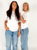 To kvinder i hvide T-shirts og blå jeans står sammen mod en hvid baggrund. De bærer smykker og udstråler selvsikkerhed.