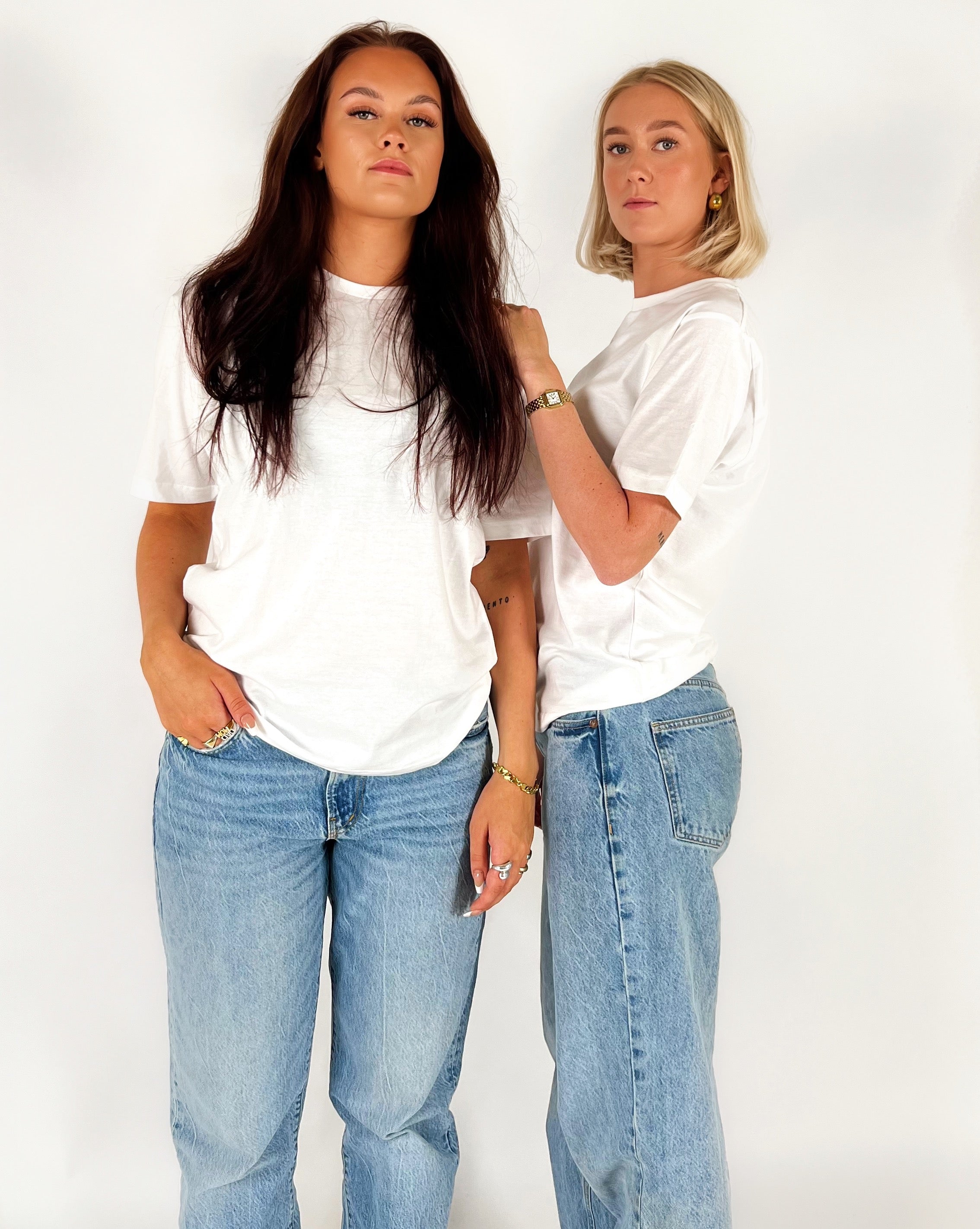 To kvinder i hvide T-shirts og blå jeans står sammen mod en hvid baggrund. De bærer smykker og udstråler selvsikkerhed.