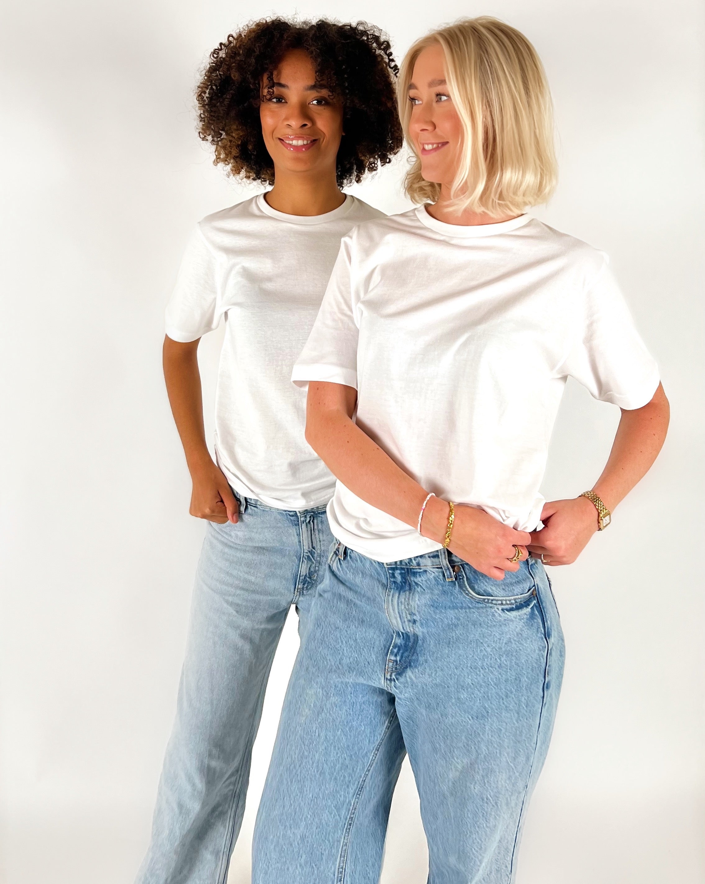 To kvinder iført hvide T-shirts og blå jeans står ved siden af hinanden og smiler i et lyst studiemiljø.