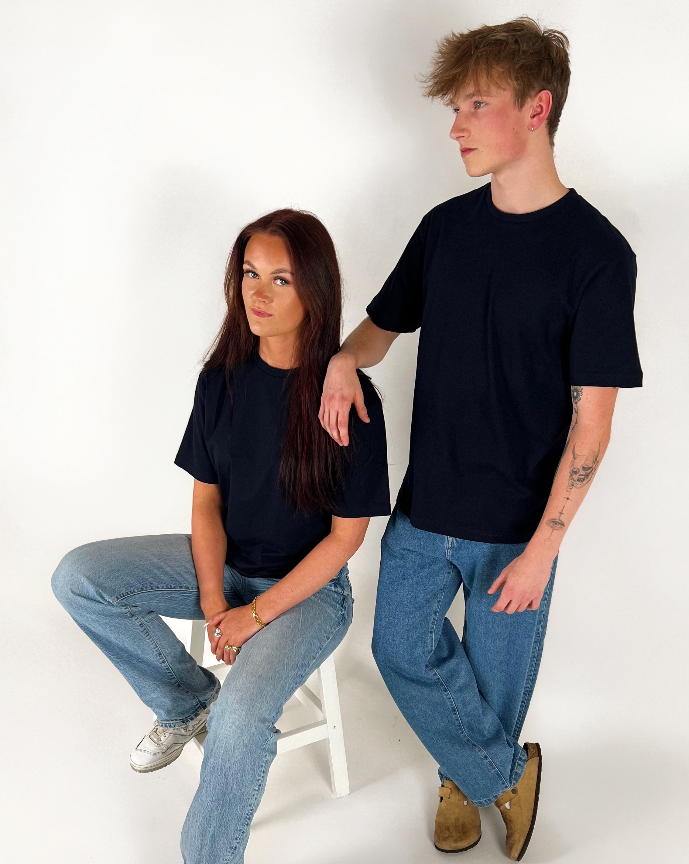 En person sidder på en hvid stol, iført en sort t-shirt og jeans, mens en anden person står ved siden af og hviler en arm på stolens ryglæn. Begge er i et lyst rum.