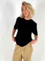 Person i sort t-shirt står med hænderne i lommerne, iført lyse bukser, med hvid baggrund.