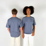 To personer står ved siden af hinanden; en vender ryggen til, mens den anden ser fremad. De bærer begge grå t-shirts og hvide bukser, i et neutralt rum.