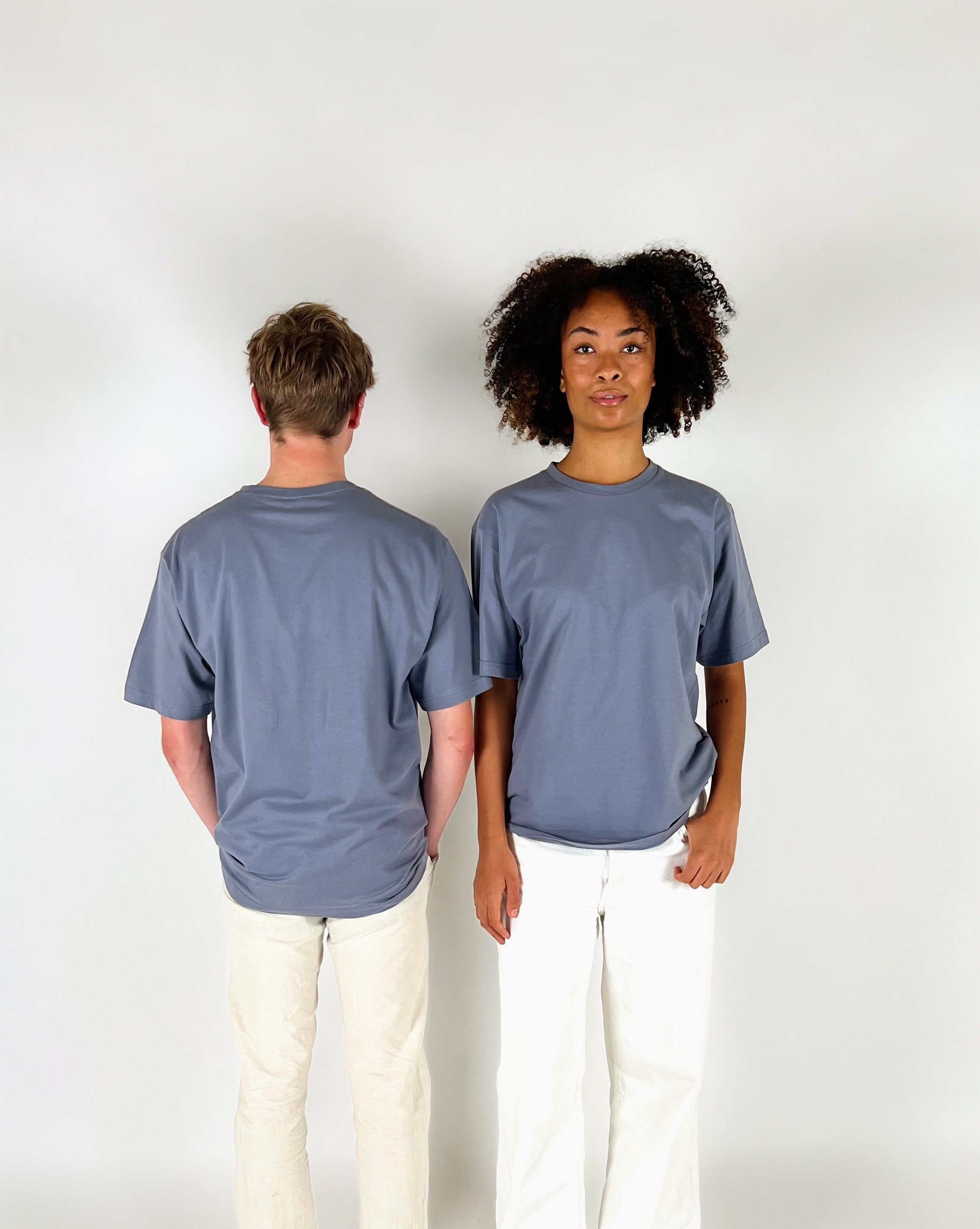 To personer står ved siden af hinanden; en vender ryggen til, mens den anden ser fremad. De bærer begge grå t-shirts og hvide bukser, i et neutralt rum.