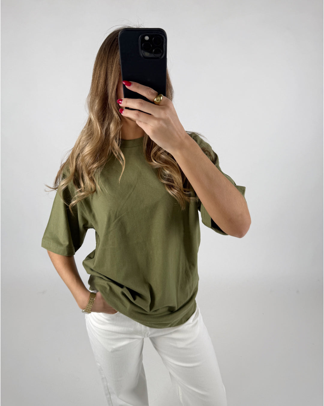 En person iført en grøn T-shirt og hvide bukser holder en sort mobiltelefon foran deres ansigt. Baggrunden er lys og neutral.