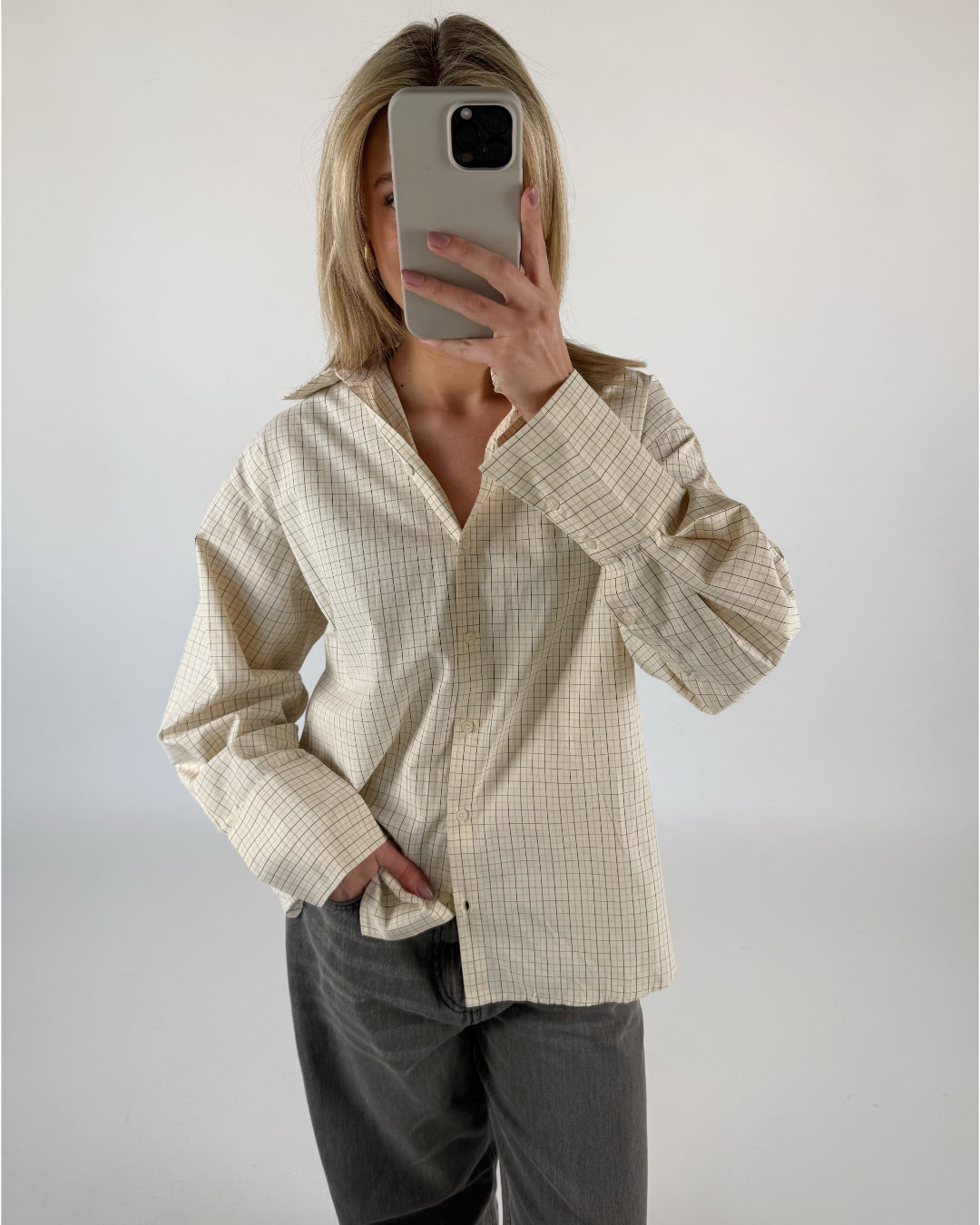 Person tager selfie, holder telefon op foran ansigtet. Iført lysegrøn ternet skjorte og grå jeans. Baggrund er minimalistisk og lys.