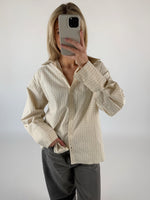 Person tager selfie, holder telefon op foran ansigtet. Iført lysegrøn ternet skjorte og grå jeans. Baggrund er minimalistisk og lys.