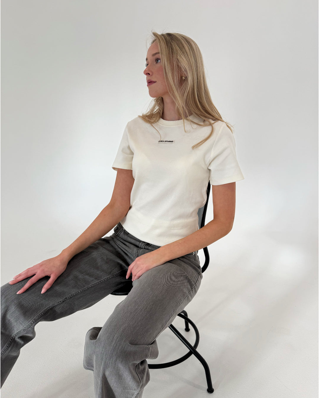 En person sidder på en stol iført en hvid t-shirt med teksten "ASOS DESIGN" og grå jeans. Baggrunden er en simpel, lys væg.
