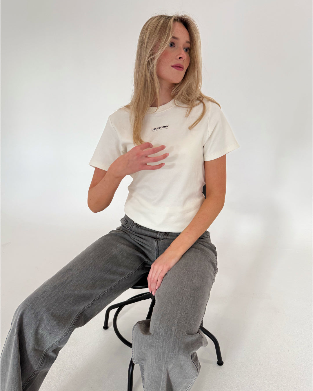 Kvinde sidder på en skammel, hånd på brystet, iført en hvid t-shirt med teksten "Axel Studio" i et lysegråt rum.
