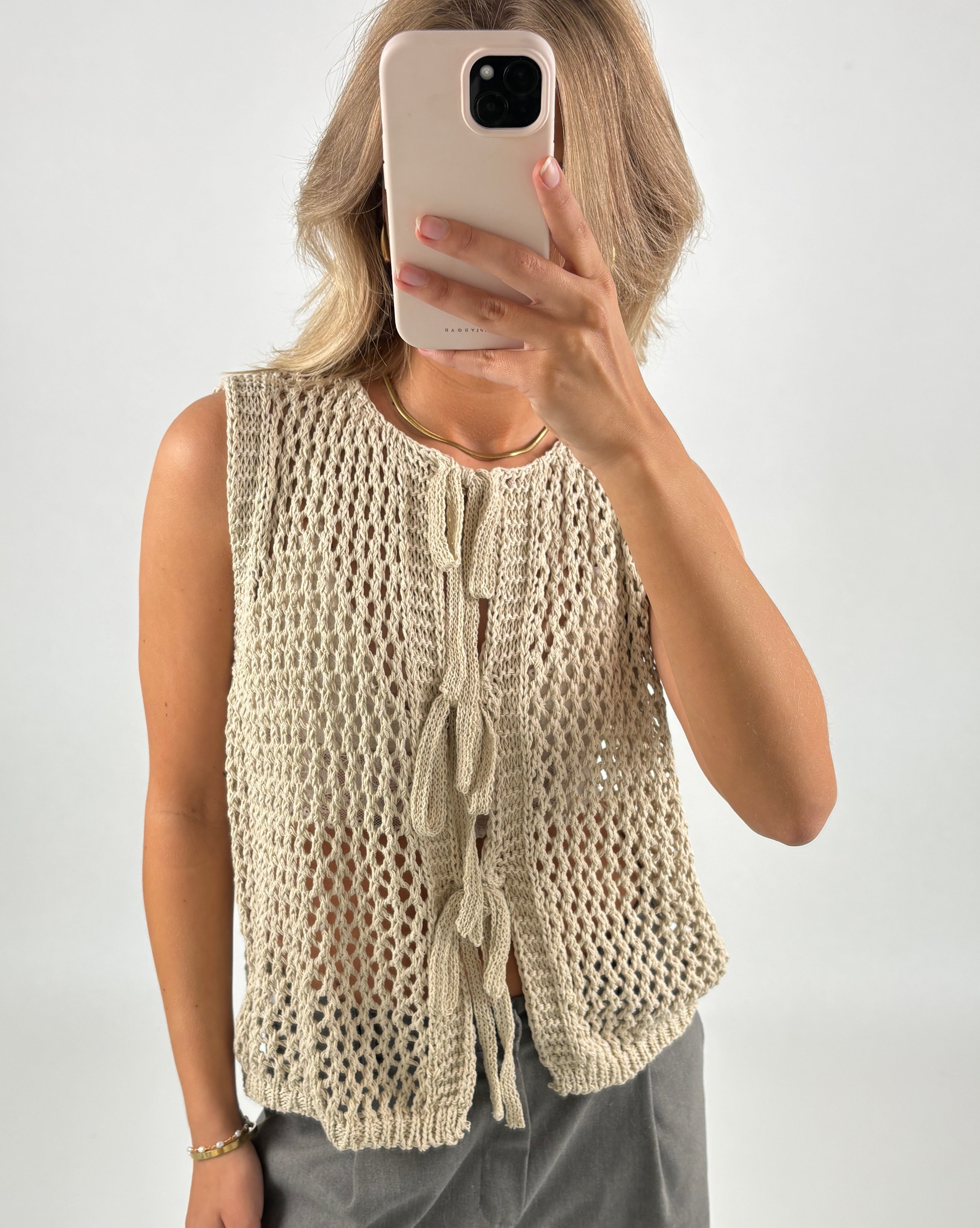 En person iført en beige, strikket vest tager et billede med en smartphone. Personen står i et lysegråt område og holder telefonen op foran ansigtet.
