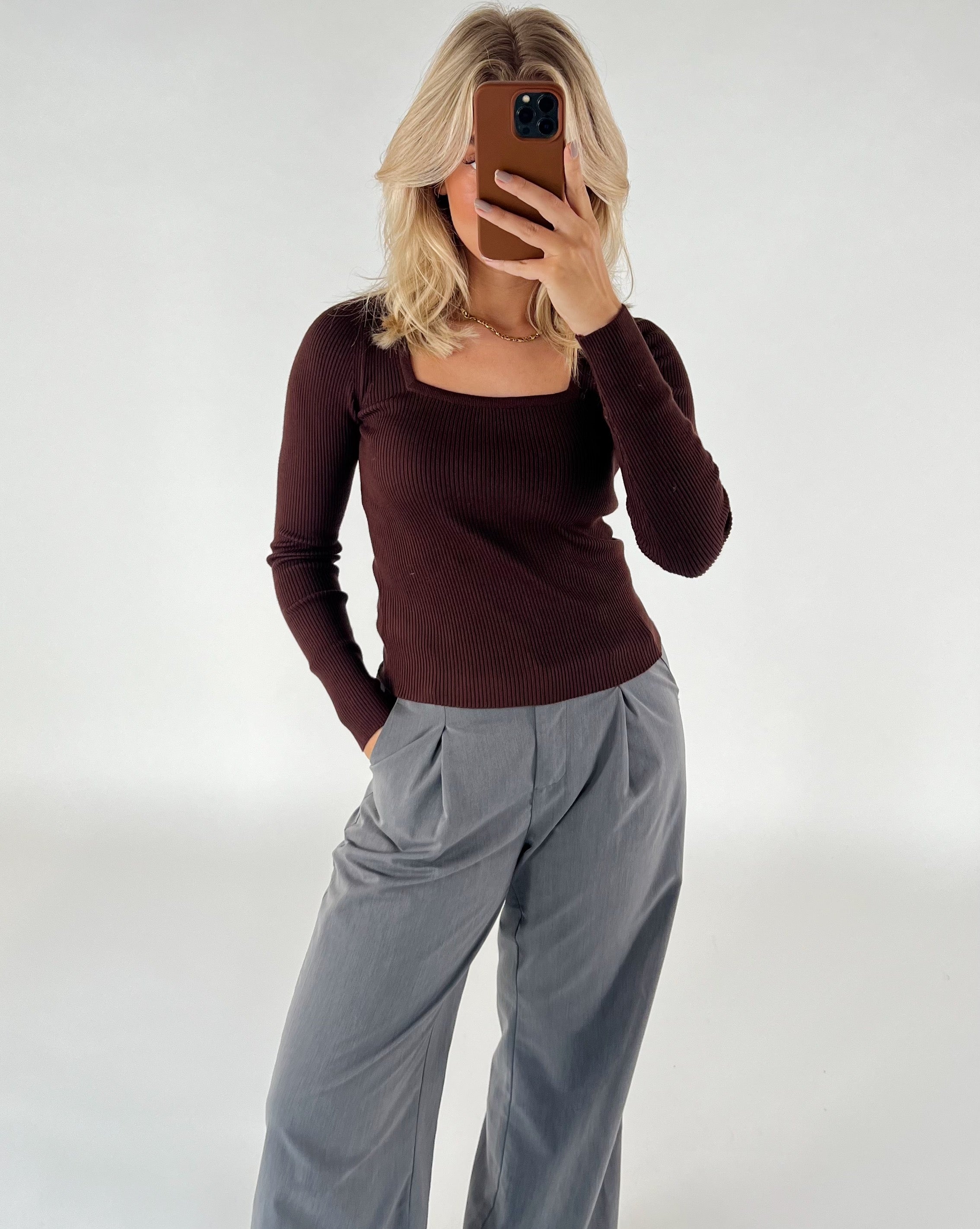 Person tager et spejl-selfie, iført brun bluse og grå bukser, i et neutralt oplyst rum. Personen dækker ansigtet med en telefon og har hånden i lommen på bukserne.