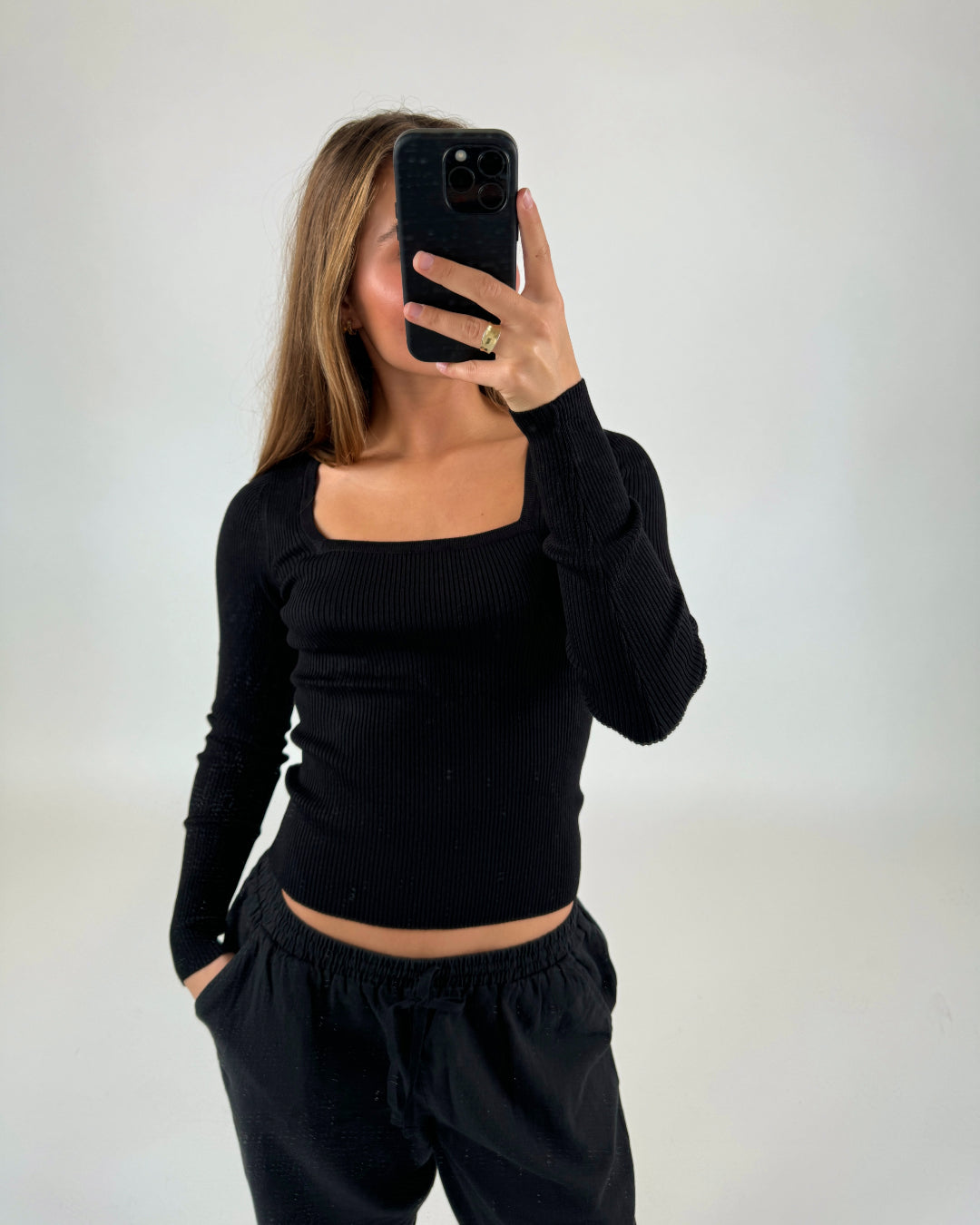 En person iført sort langærmet t-shirt og bukser tager et spejlbillede med en smartphone i en minimalistisk baggrund. Hånden er tæt på kameraet, og hovedet er tildækket.
