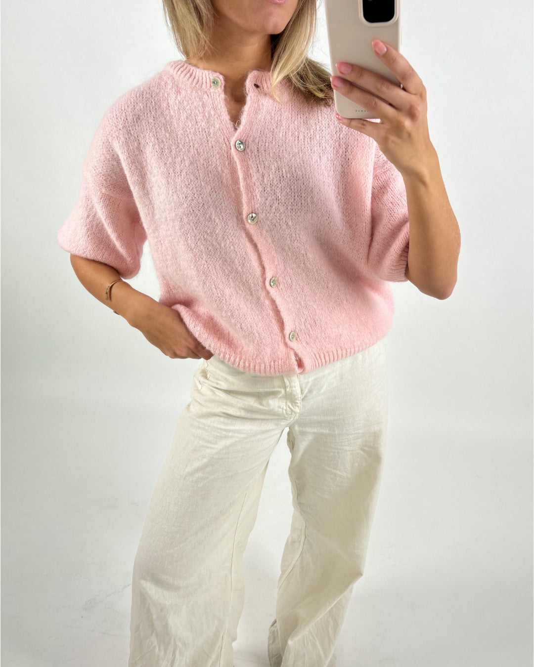 Kvinde iført pink cardigan og hvide bukser, tager selfie med smartphone i neutralt lys. Baggrunden er enkel og hvid. Ingen tekst til stede.