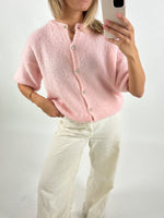 Kvinde iført pink cardigan og hvide bukser, tager selfie med smartphone i neutralt lys. Baggrunden er enkel og hvid. Ingen tekst til stede.
