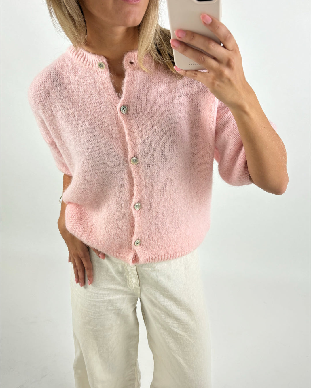 Person i en lyserød cardigan knapper med hvide bukser holder en smartphone. Stående mod en hvid baggrund i en lys, neutral indendørs setting.