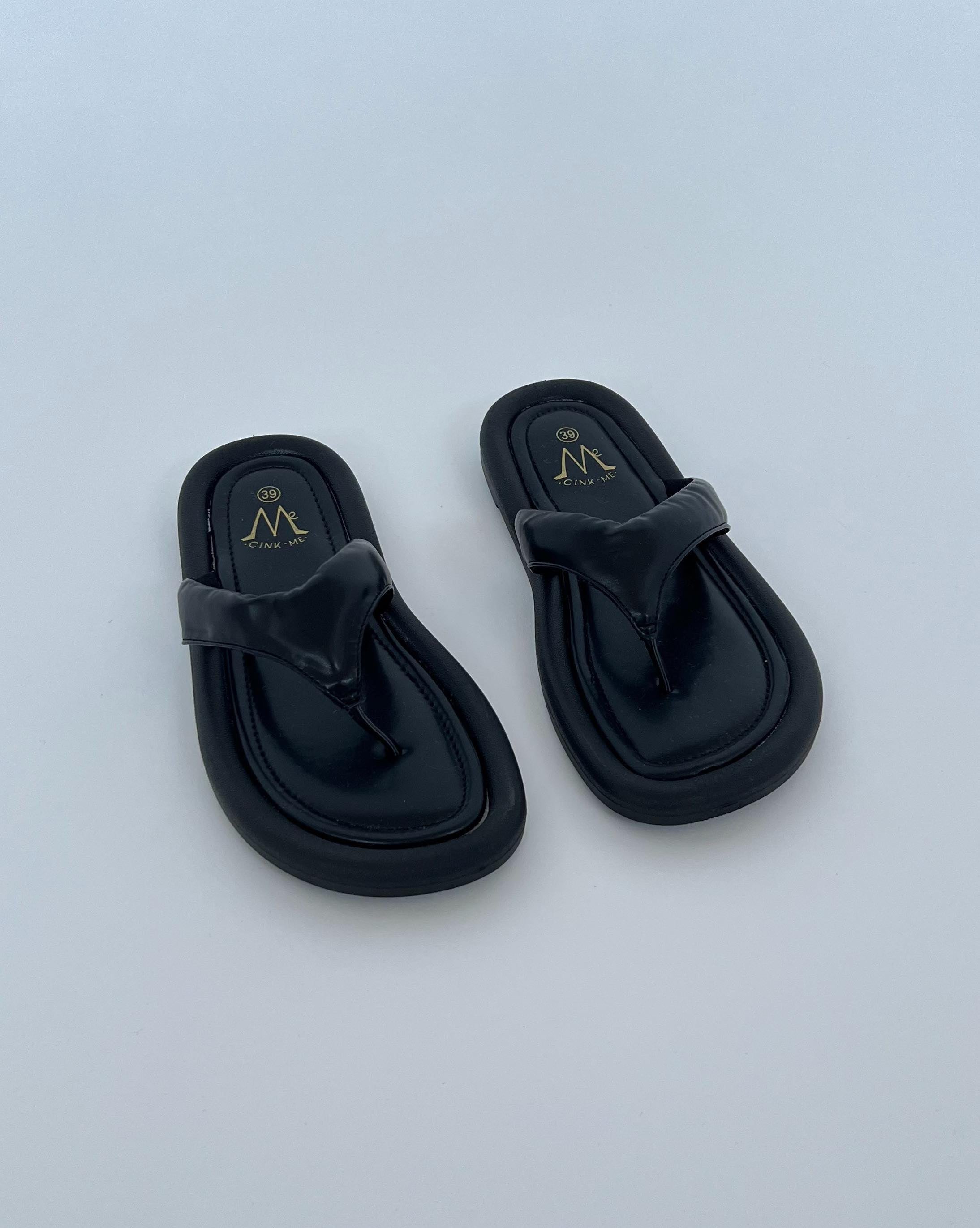 Sorte sandaler ligger stille på en lys baggrund. Sandalerne har en skinnende finish med logoet "Me CINK ME" trykt i guld på indersålerne.
