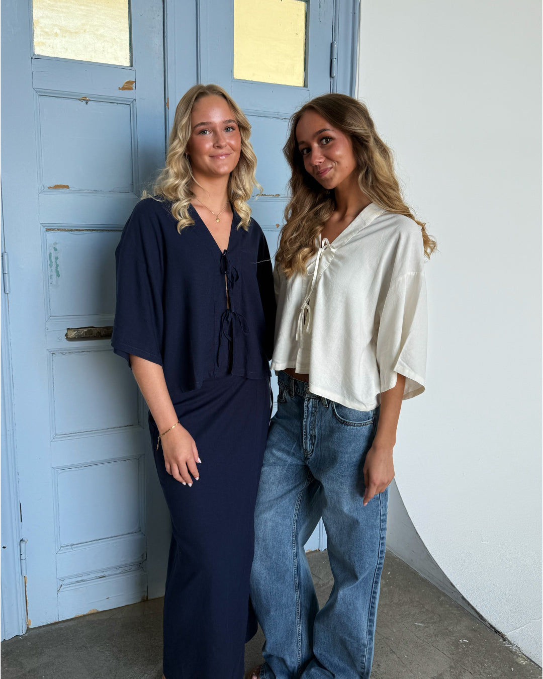 To unge kvinder står sammen og smiler foran en lyseblå dør med glasruder. Den ene bærer en mørk kjole, den anden en lys bluse og jeans.