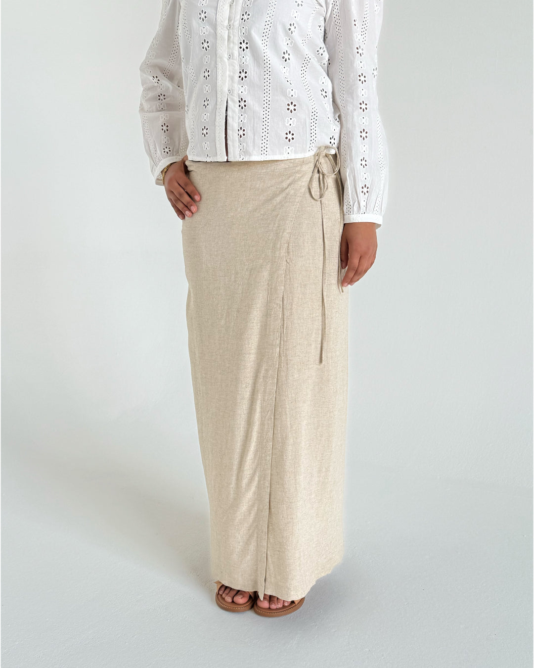 En person iført en hvid, blomstermønstret, langærmet bluse og en lang, beige nederdel står i et minimalistisk rum med en lys baggrund.