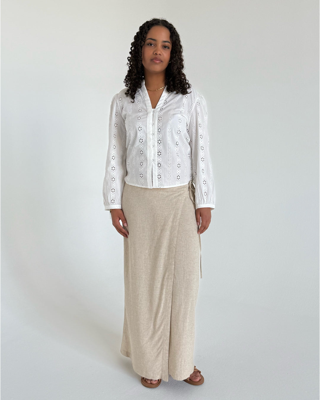 En person står iført en hvid bluse med broderede mønstre og en lang beige nederdel. De har brunt, krøllet hår og står i et neutralt, indendørs rum med en lys baggrund.