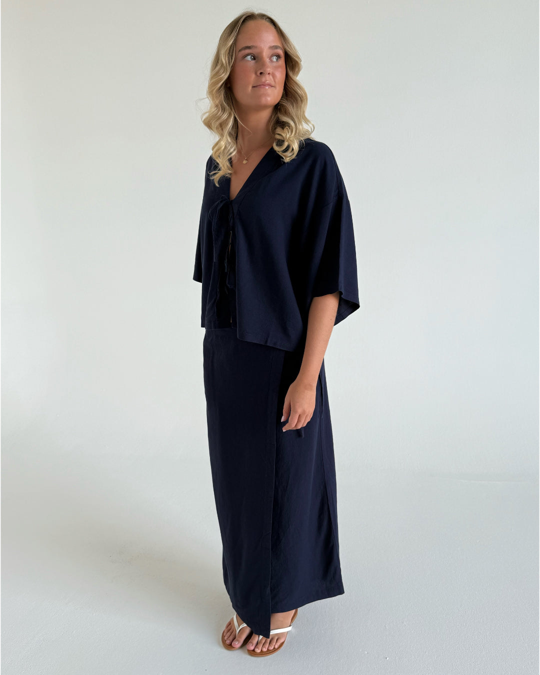 En person iført en mørkeblå kjole og sandaler står og kigger væk i et minimalistisk, lyst område.