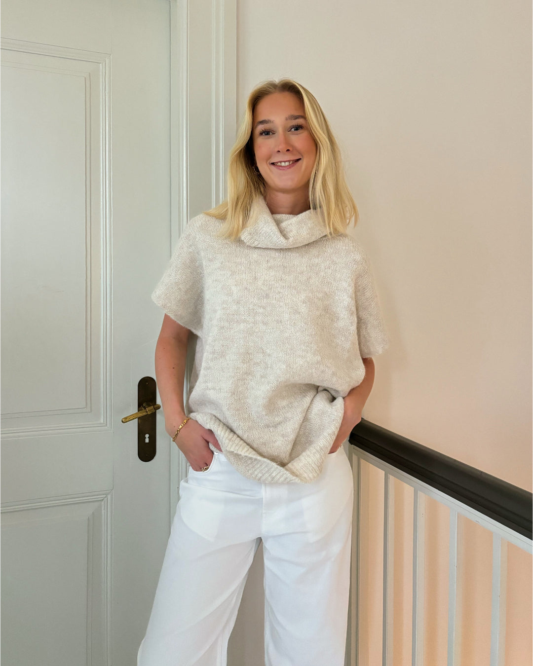En person står og smiler iført en beige sweater og hvide bukser. Personen læner sig mod en lys væg ved siden af en dør.