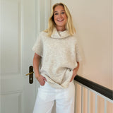 En person står og smiler iført en beige sweater og hvide bukser. Personen læner sig mod en lys væg ved siden af en dør.