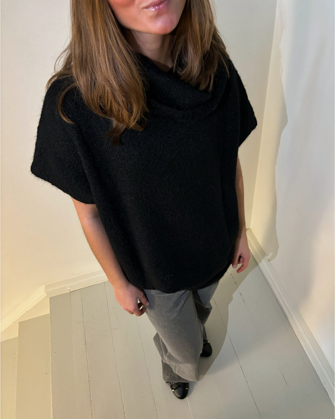En person iført sort sweater står på en gråmalet trappe. Personen er delvist uden for billedet, med langt brunt hår og grå bukser. Væggen er hvid baggrund.