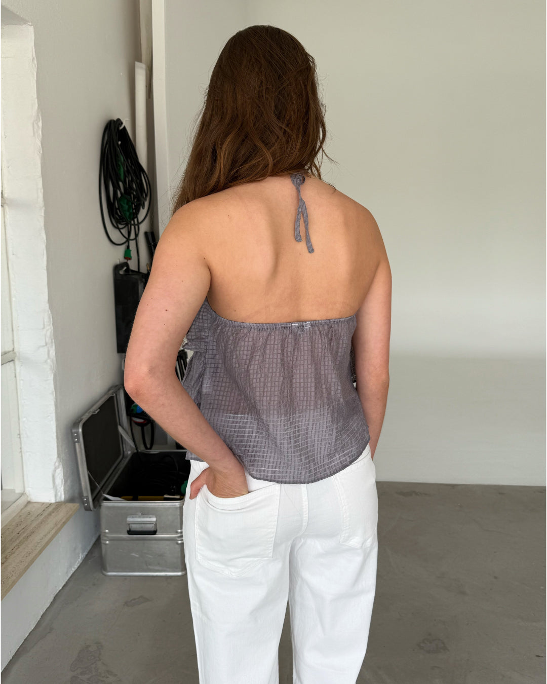 Modelfoto af pige som bærer en grå BRITNEY halterneck top med blomst på bryst, samt hvide jeans. Set bagfra. Alt er fra BySofieSønderby
