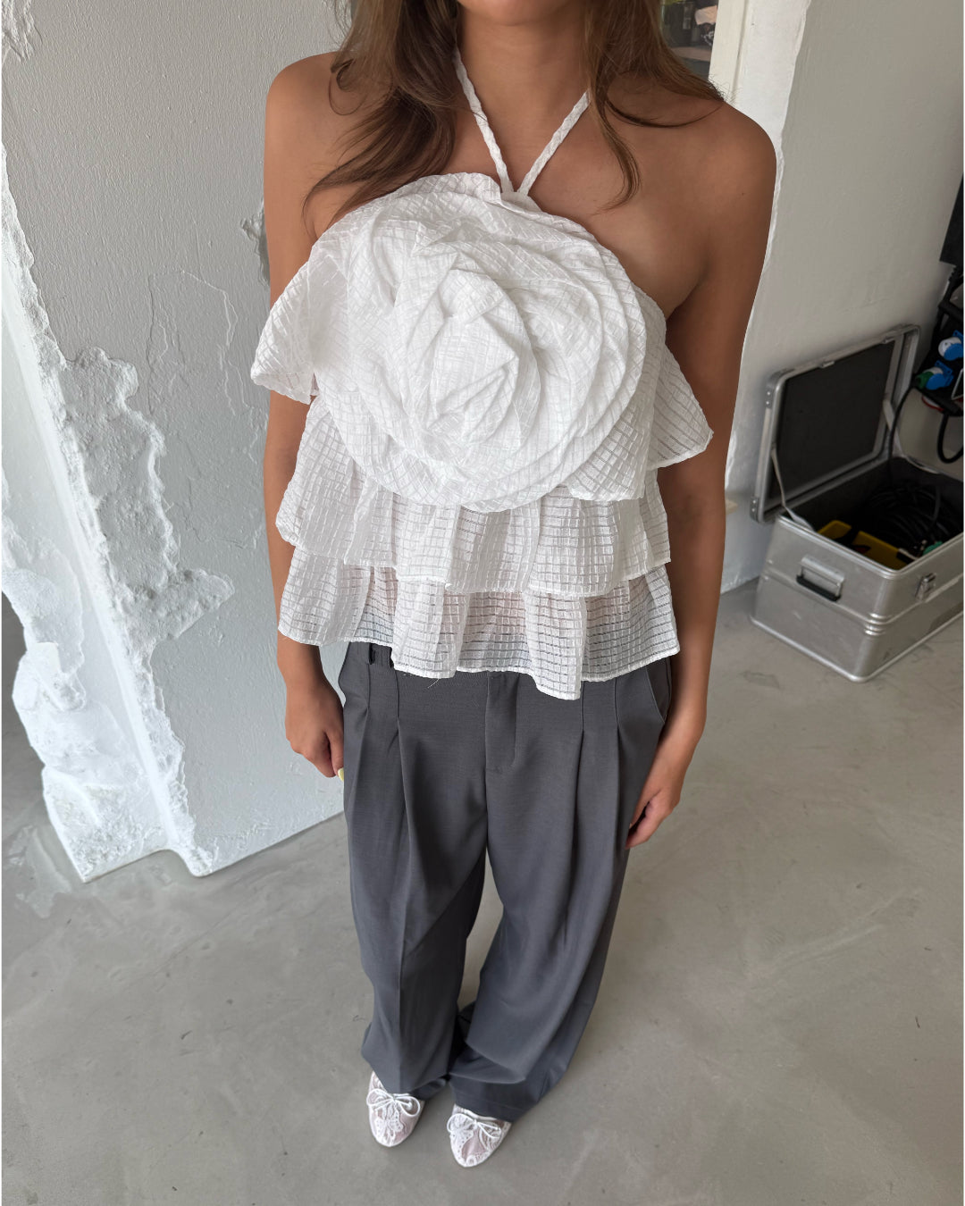 Modelfoto af pige som bærer en hvid BRITNEY halterneck top med blomst på bryst, samt grå kontorbukser og hvide ballerinaer. Alt er fra BySofieSønderby