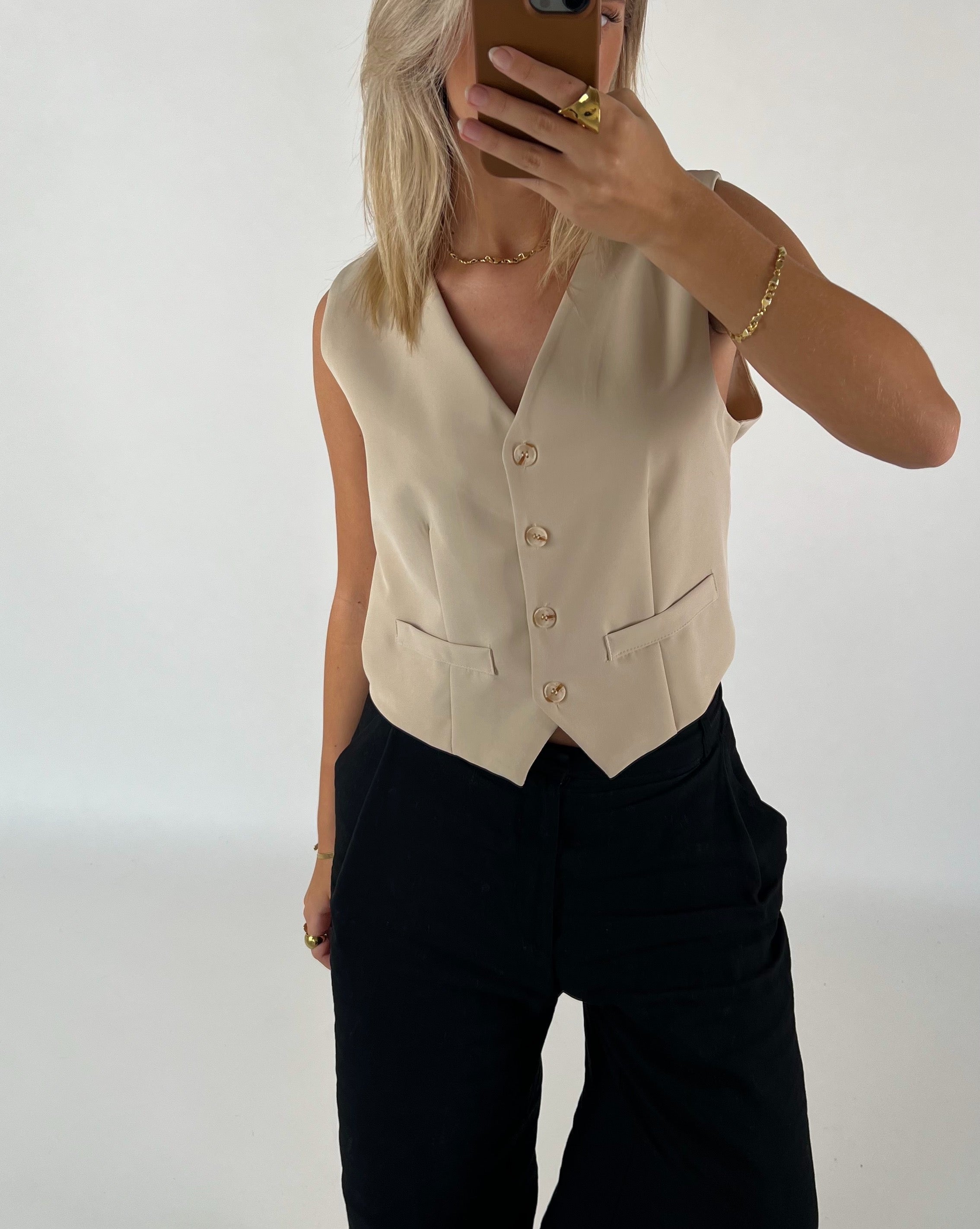 En person iført en beige vest tager et selfie med en smartphone. Personen har blondt hår og bærer sorte bukser og guldsmykker i et minimalistisk, lyst rum.