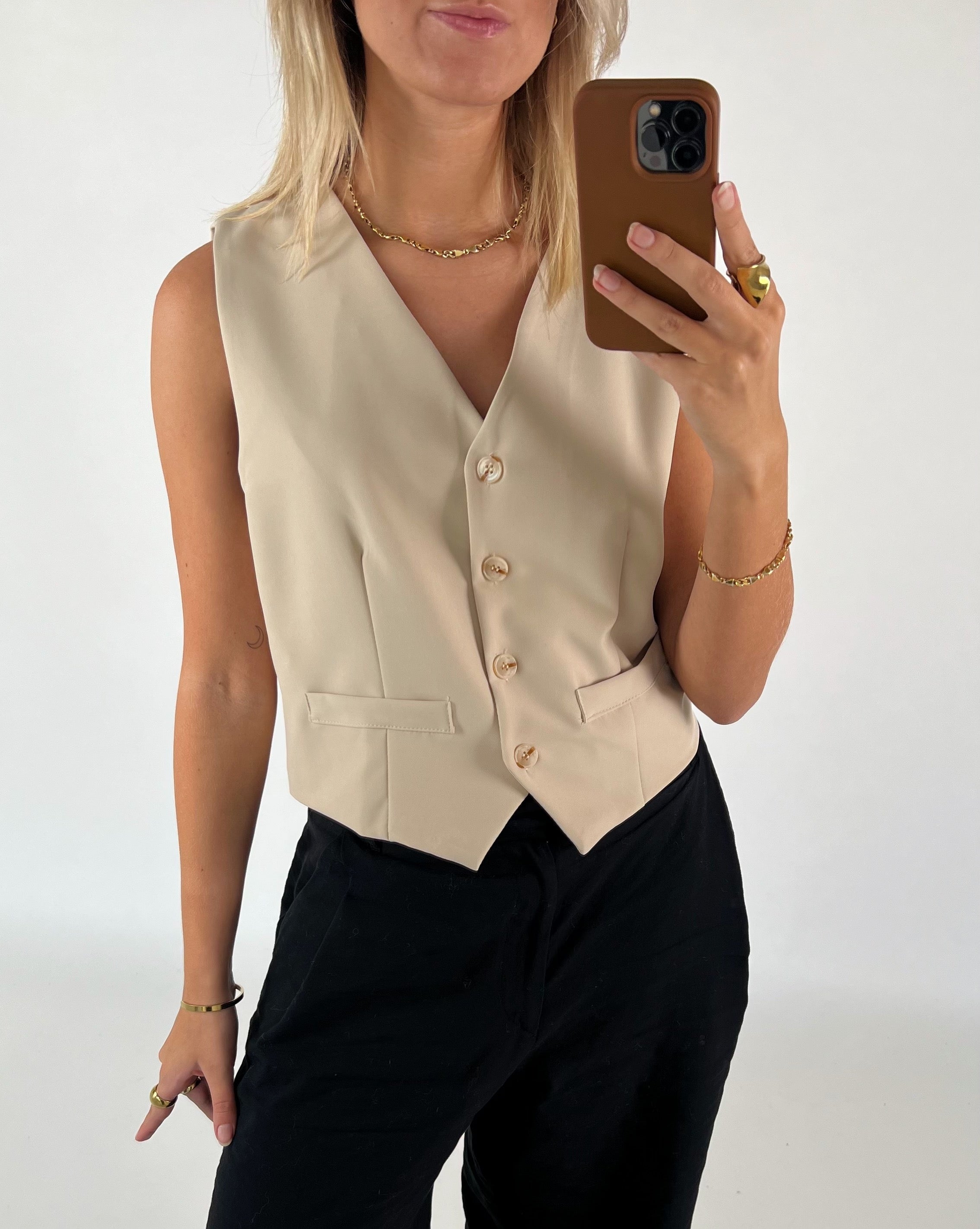 En person iført en beige vest tager en selfie med en brun mobil i et lyst rum. Personen bærer også sorte bukser og gyldent smykke.