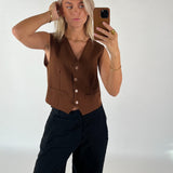 Person tager en selfie med en smartphone, iført en brun vest og sorte bukser, i et lys oplyst rum. Personen har blondt hår og ser direkte ind i kameraet.