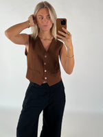 Person tager en selfie med en smartphone, iført en brun vest og sorte bukser, i et lys oplyst rum. Personen har blondt hår og ser direkte ind i kameraet.