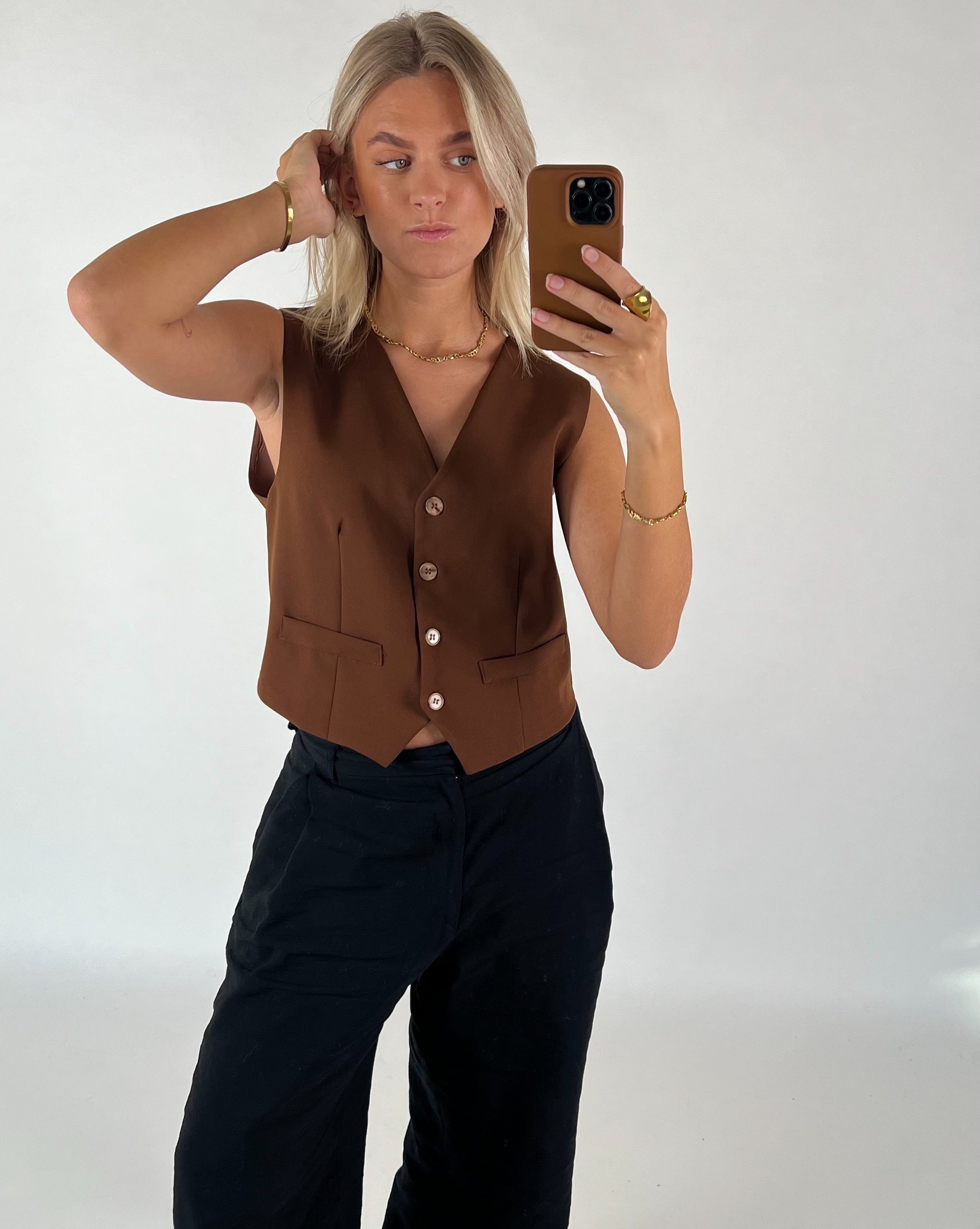 Person tager en selfie med en smartphone, iført en brun vest og sorte bukser, i et lys oplyst rum. Personen har blondt hår og ser direkte ind i kameraet.