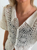En hvid hæklet cardigan bæres af en person. Trøjen er knapper foran og har et blomstermønster. Personen har en guldhalskæde på, i et lyst indendørs miljø.