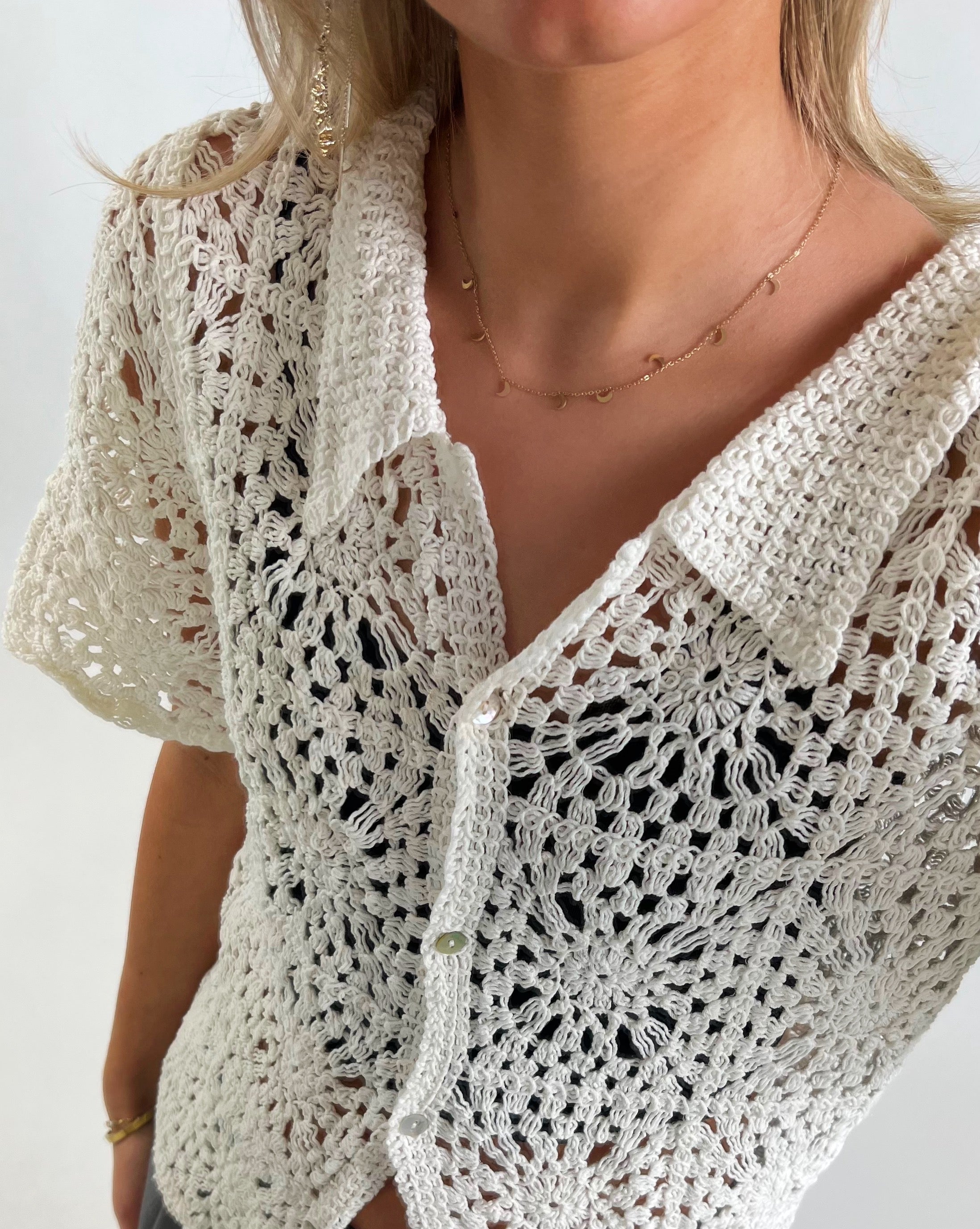 En hvid hæklet cardigan bæres af en person. Trøjen er knapper foran og har et blomstermønster. Personen har en guldhalskæde på, i et lyst indendørs miljø.