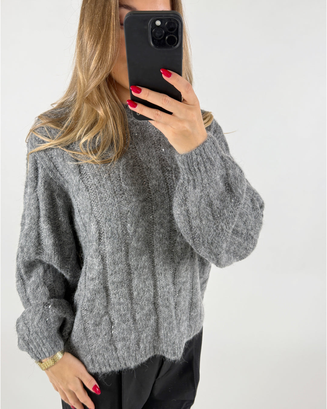 En person iført en grå striktrøje tager et selfie med en sort telefon i hånden. Personen befinder sig foran en neutral, hvid baggrund.