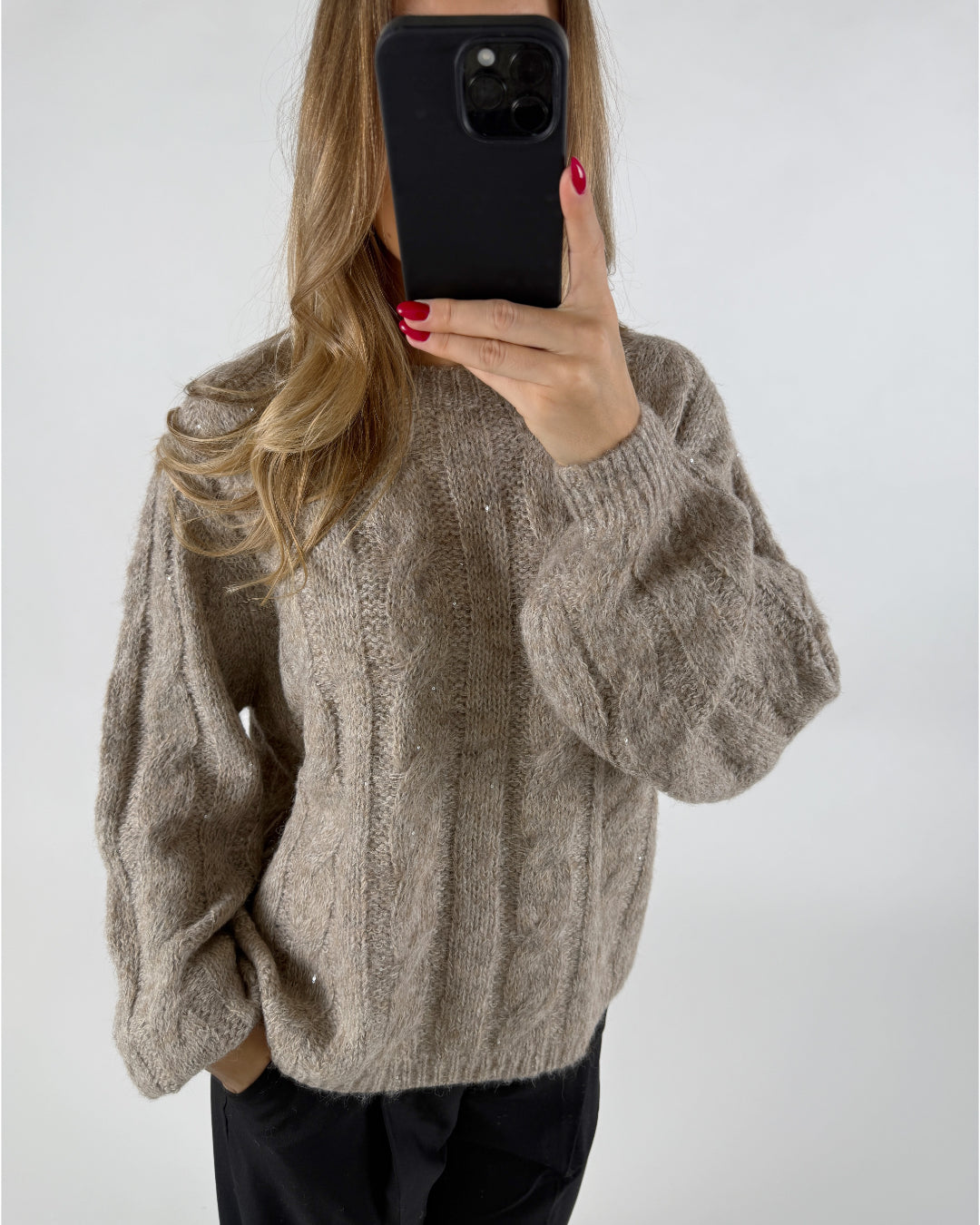 Person holder en telefon foran ansigtet, iklædt en strikket beige sweater med kabelmønster, i en neutral indendørs setting. Personen har langt, bølget hår og rødmalede negle.