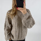 Person holder en telefon foran ansigtet, iklædt en strikket beige sweater med kabelmønster, i en neutral indendørs setting. Personen har langt, bølget hår og rødmalede negle.