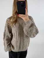 Person holder en telefon foran ansigtet, iklædt en strikket beige sweater med kabelmønster, i en neutral indendørs setting. Personen har langt, bølget hår og rødmalede negle.