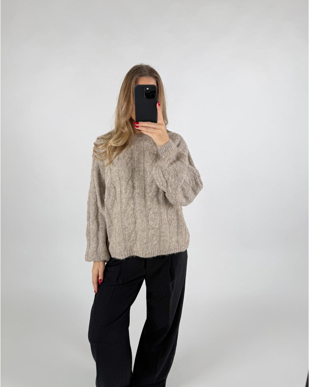 En person iført en beige striktrøje tager et spejlbillede med en smartphone. Personen står i et neutralt rum med en enkel baggrund.