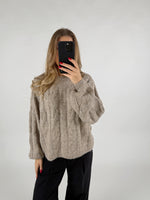 En person iført en beige striktrøje tager et billede af sig selv med en smartphone. Personen står i et rum med en neutral, lys baggrund.