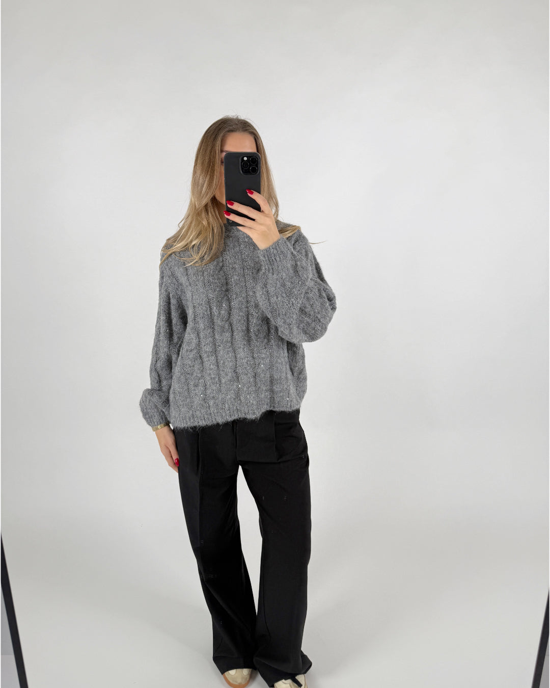 Person tager et spejlbillede iført en grå striktrøje og sorte bukser. Personen holder en telefon med rødnegler i et minimalistisk, lyst rum.