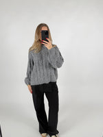 Person tager et spejlbillede iført en grå striktrøje og sorte bukser. Personen holder en telefon med rødnegler i et minimalistisk, lyst rum.