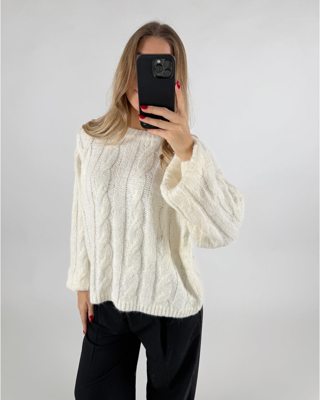 En person iført en hvid strikket sweater tager et spejlfoto med en sort telefon i hånden, med en ensfarvet lys baggrund.