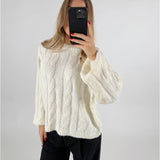 En person iført en hvid strikket sweater tager et spejlfoto med en sort telefon i hånden, med en ensfarvet lys baggrund.