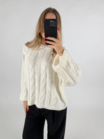 En person iført en hvid strikket sweater tager et spejlfoto med en sort telefon i hånden, med en ensfarvet lys baggrund.