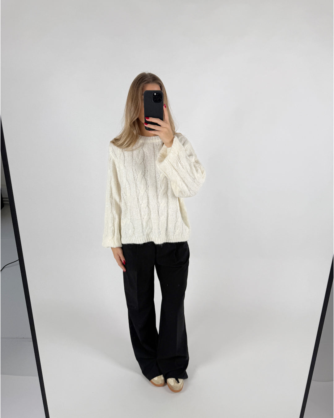 En person iført en hvid sweater og sorte bukser tager et selfie foran et spejl i et minimalistisk rum med hvid baggrund.
