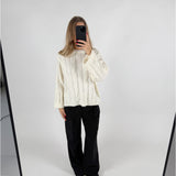 En person iført en hvid sweater og sorte bukser tager et selfie foran et spejl i et minimalistisk rum med hvid baggrund.