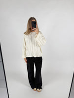 En person iført en hvid sweater og sorte bukser tager et selfie foran et spejl i et minimalistisk rum med hvid baggrund.