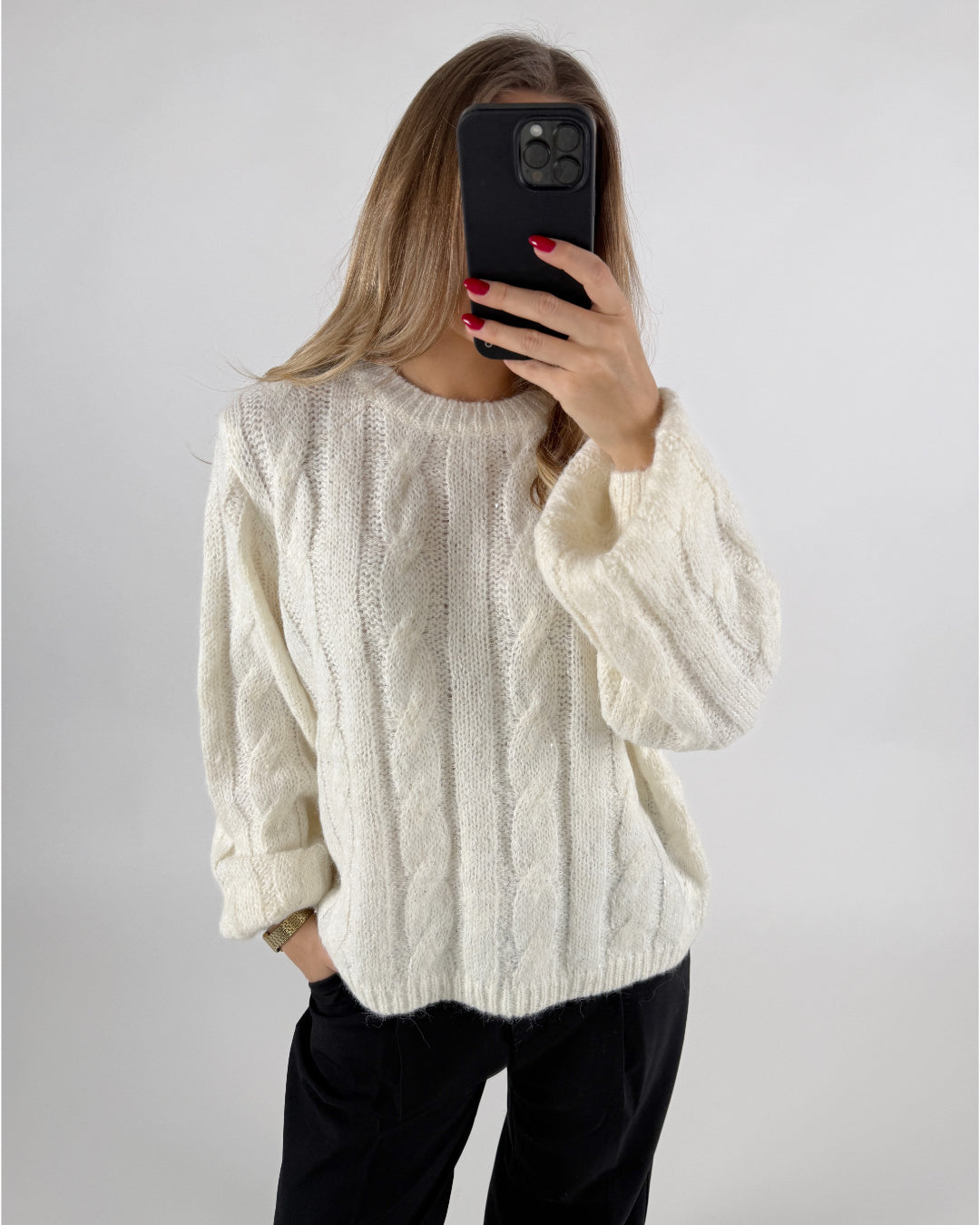 Kvinde iført hvid sweater tager selfie med telefon foran ansigt; langærmet trøje med flettemønster. Hendes negle er malet rød, og hun står mod en neutral baggrund.